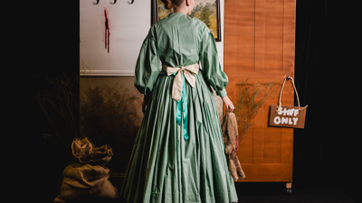 Woman in long green dress with a white apron, sitting in front of a wooden backdrop with a framed painting, hanging animal carcass, dried plants, and a "Staff Only" sign. 