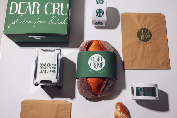 Flat lay of 'Dear Crumb' branding materials including green packaging box, brown paper bags, branded stickers, a napkin, and a loaf of bread with a green paper banded wrap.