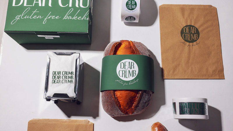 Flat lay of 'Dear Crumb' branding materials including green packaging box, brown paper bags, branded stickers, a napkin, and a loaf of bread with a green paper banded wrap.