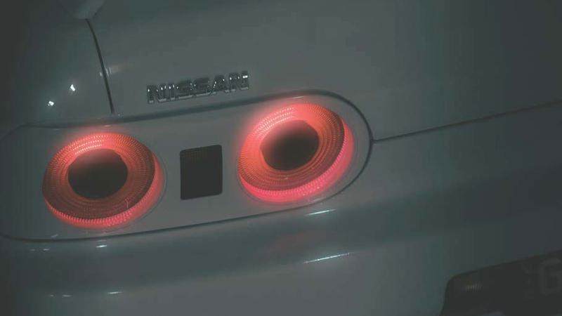 Close-up of a light-coloured Nissan R32 Skyline car's rear panel, showing two illuminated, circular red tail lights, photographed in low lighting.