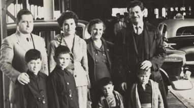 Extended family standing around suitcase on Station Pier, under a large roofed structure. Two men, two women, two boys and two infant children. There is a large suitcase in the foreground with rugs strapped to the side. Cars are parked in the background.