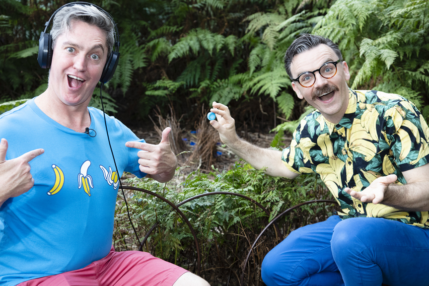 The Listies, comic duo Richard Higgins and Matt Kelly in Forest Gallery promoting their Melbourne Museum Audio Guide App. Matt is wearing headphones, Richard is holding blue bottle cap and pointing to Matt's blure t-shirt.