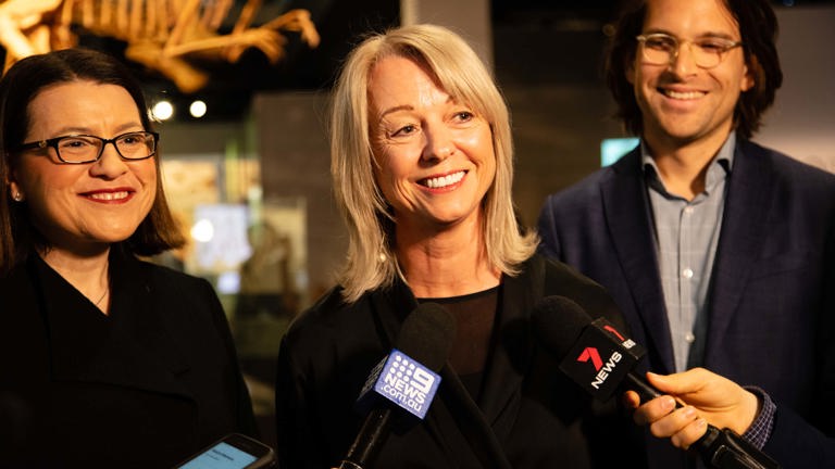 A woman speaking in to news microphones. A woman stands to her left and a man to her right