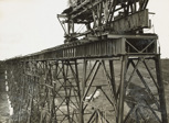A travelling crane is on top of one of the towers of the Maribyrnong River Viaduct on the Albion to Broadmeadows line, Keilor East, 1928. A girder, awaiting placement, is sitting on a cradle beneath it.