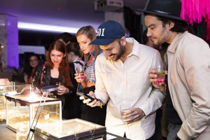 People holding drinks peering in display boxes. One person is taking a photo with a phone
