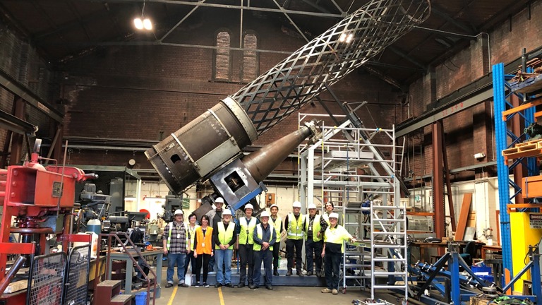 Group of people wearing hi-vis vest stand in front of a large metal cylinder in an engineering workshop