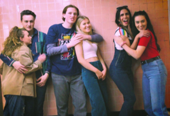 Still of six people standing against a tiled wall, hugging each other in pairs