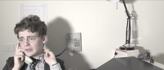 Still of a boy that is visibly stressed, speaking on a telephone next to a desk