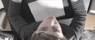Still looking down on a boy sitting at a desk drawing on a piece of paper