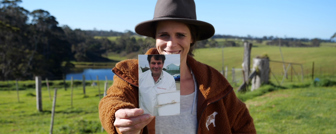 Still of a woman in a wide-brim hat smiling and holding up a photograph of a man 