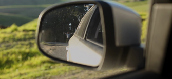 Film still showing a close up of the mirror on the passenger side of a car, in which a figure can be seen waving in the distance behind the vehicle