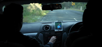 Film still showing two men in the front of a car.  A photo of a woman and child is taped to the dashboard