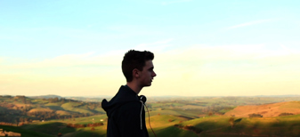 Film still showing the side profile of a young man, with headphones around his neck, standing above an expanse of rolling green hills in the evening sun