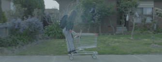 Film still shows the side profile of a girl riding on the back of a shopping trolley down the street outside suburban homes