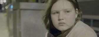 Film still shows a close up of a melancholic girl, under the white lights of a train station at night time 