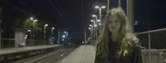 Film still shows a young girl with long brown hair stands alone, looking troubled, at a train station at night time