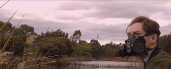 Close up of a concerned young man wearing a gas mask sitting by a river