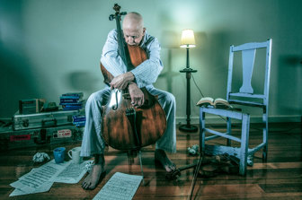 A seated older man is dressed in his pyjamas while sadly holding a cello, with his chin resting on the top of the instrument, looking contemplatively at his sheet music on the ground. Books, music, and mugs are strewn around him. The photograph comprises mainly blue/grey objects, except for the cello, and an overall blue colour pallete