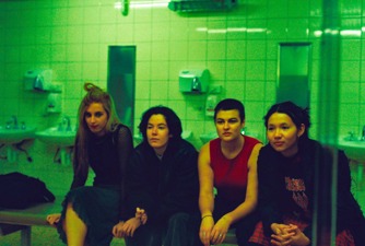 Four girls dressed in punk clothes are sitting in a row on a bench  a green-lit, tiled public bathroom. 