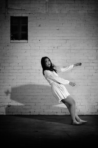 A black and white photograph of a fearful girl with long black hair, wearing a short white dress,  falling backwards, towards a shadow of a coffin on the ground, as if she is being drawn towards it