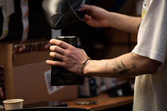 A close up of a tattooed arm puring ground coffee into black packaging