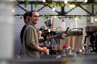 A young man making coffee at a coffee machine smiles in the direction of the photographer, seemingly unaware of the image being taken