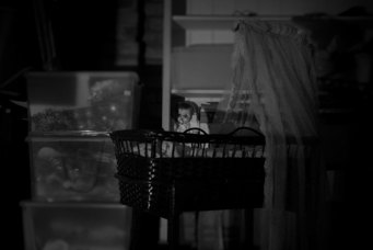 A black and white photo of an old-fashioned wicker bassinet with a lace canopy. An old-fashioned doll sits upright, with her arm upwards. 