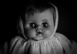 A black and white photo of an old-fashioned plastic doll dressed in a woolen jumper and hood. 
