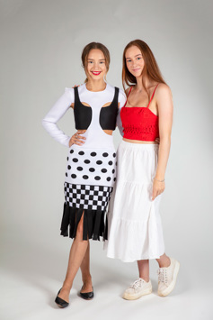 A girl is modelling a long-sleeved black and white dress with cutouts. The dress has a black crop top beneath. A girl wearing a red top and white skirt has her arm around the model 