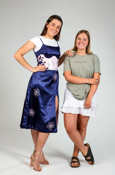 A girl with brown hair models a blue satin dress, worn over a white t-shirt. A pink resin brace is work over the dresss. A shorter blond girl stands to the right, smiling at the camera. 