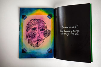 A photograph of an open art zine showing two pages, one is a colourful page with a hand-drawn illustration of a face on transparent acetate, the other is a black page with white text, in a hand-written style