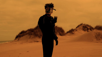 A man wearing black protective gear, including gloves and a gas mask, holds up a black box while standing on an empty beach. The light is dim, yellow and hazy