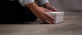 A close up of a pair of hands placed either side of a white box that is sitting on a concrete floor. The person is kneeling and their sleeves are rolled up to reveal a white shirt. 