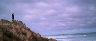 A girl dressed in a black tracksuit stands on a grassy sand dune looking out to the ocean. The weather is grey, cloudy and windy. 