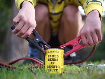 A young boy crouched on the grass, connecting two jump leads to a can of baked beans