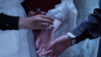 A close up of a three hands extended forward. A maid applies perfume from a glass bottle to the wrist of a woman dressed in white lace, whose hand is held by another maid. 