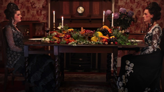 Two identical women dressed in elaborate period costumes sit opposite one another at a table laden with food. The room is dark and wallpapered and decorated with purple dahlias. 