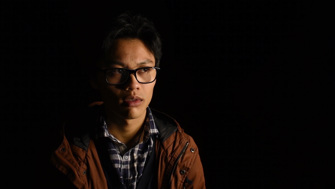 A teenage boy wearing glasses, a check shirt and a brown coat stands alone in the dark, with an apprehensive expression 