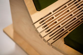 A close up of the curve in between the top and bottom of a plywood chair, showing detailed laser-cut lines and a green cushion