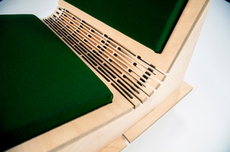 A close up of the curve in between the top and bottom of a plywood chair, showing detailed laser-cut lines and a green cushion
