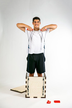 A boy demonstrates the use of a piece of exercise equipment, the 'Gym Box'.  He is standing within the box, pulling upwards on resistance bands that are attached to either side of the box.