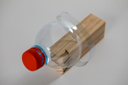 A wooden cutting tool that cuts plastic bottles. This is a small wooden block with a sharp blade embedded within it, being used to cut a spiral shape out of a plastic water bottle with a red cap is shown being spiral cut