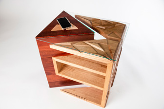 A top view of a wooden table with carved geometric details. A mobile phone is charging on top of the table, using the electrical componenents hidden within the table