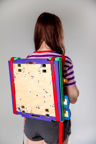A girl carries a flat-packed wooden stool, strapped to her back using red nylon straps