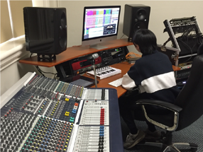 A young man at the computer in a recording studio next to a large music panel