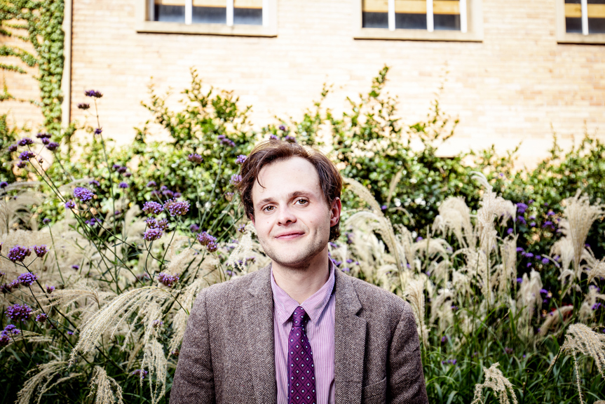 A man in a grey suit and purple shirt and tie standing in front of a garden with soft plants and a sandstone wall