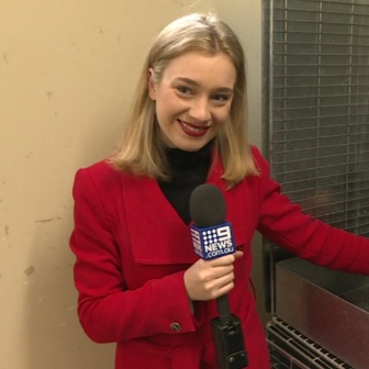 A woman with blonde hair and a red jacket stands in a corner holding a Channel 9 News microphone