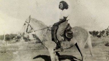 A black and white photograph of a smiling young woman atop a horse. 