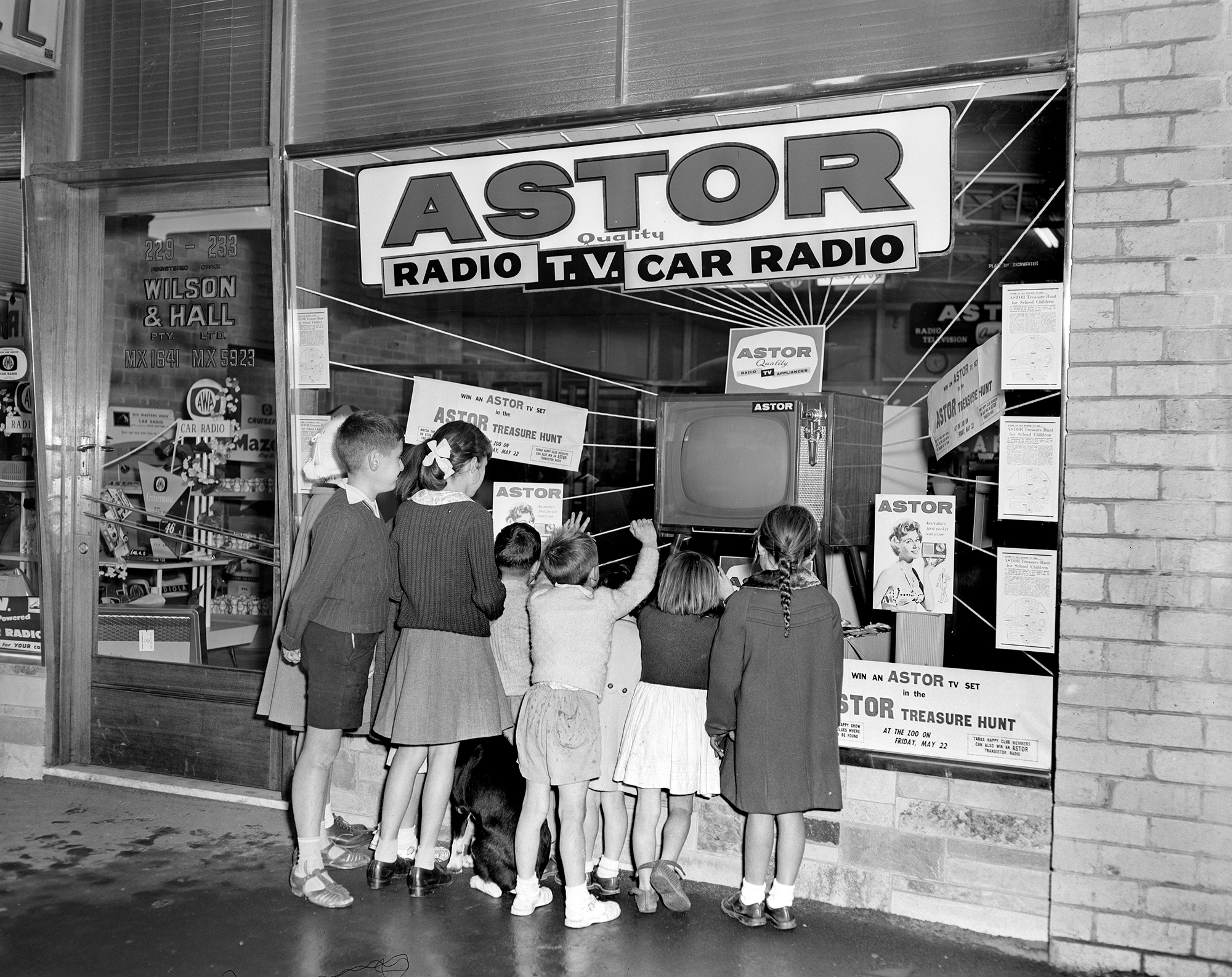 Promotional image of a group of people looking at the Wilson Electronic Store display window. There is a display of Astor radios and televisions.