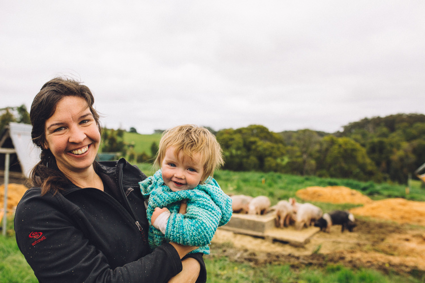 A woman beams while holding her toddler with some pigs in the background. 